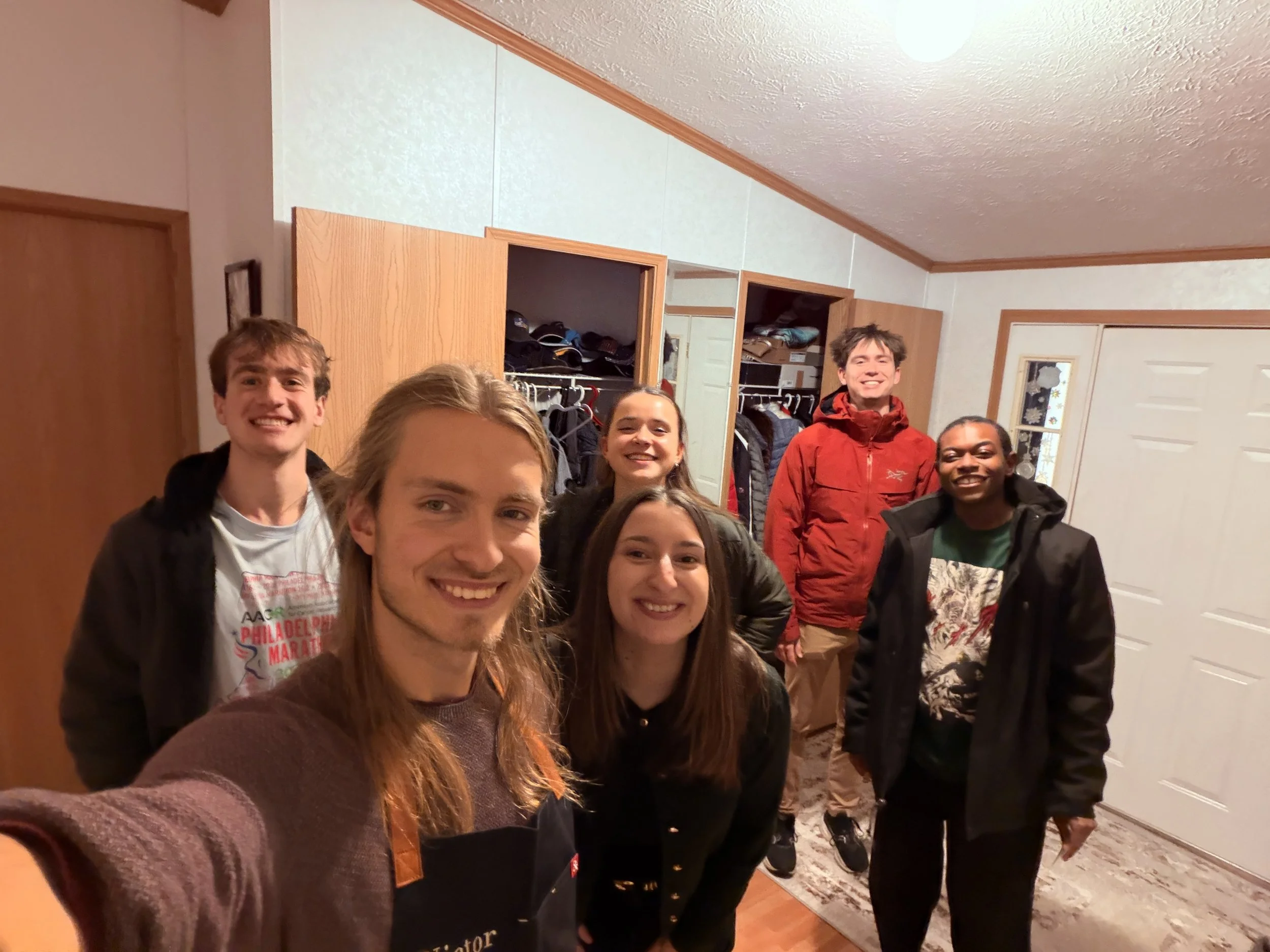 Group of smiling young adults standing together indoors, with two men and three women, in front of an open closet with clothes, and a white door with a decorative glass panel in the background.