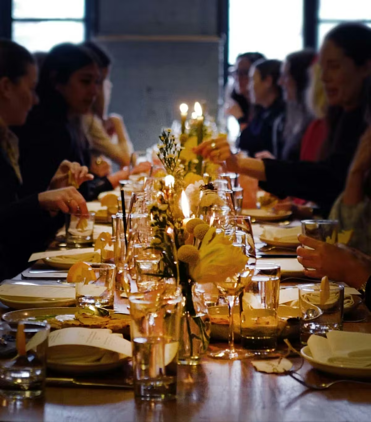 People seated at a dinner table with floral centerpieces, glasses, plates, and lit candles, engaging in conversation.