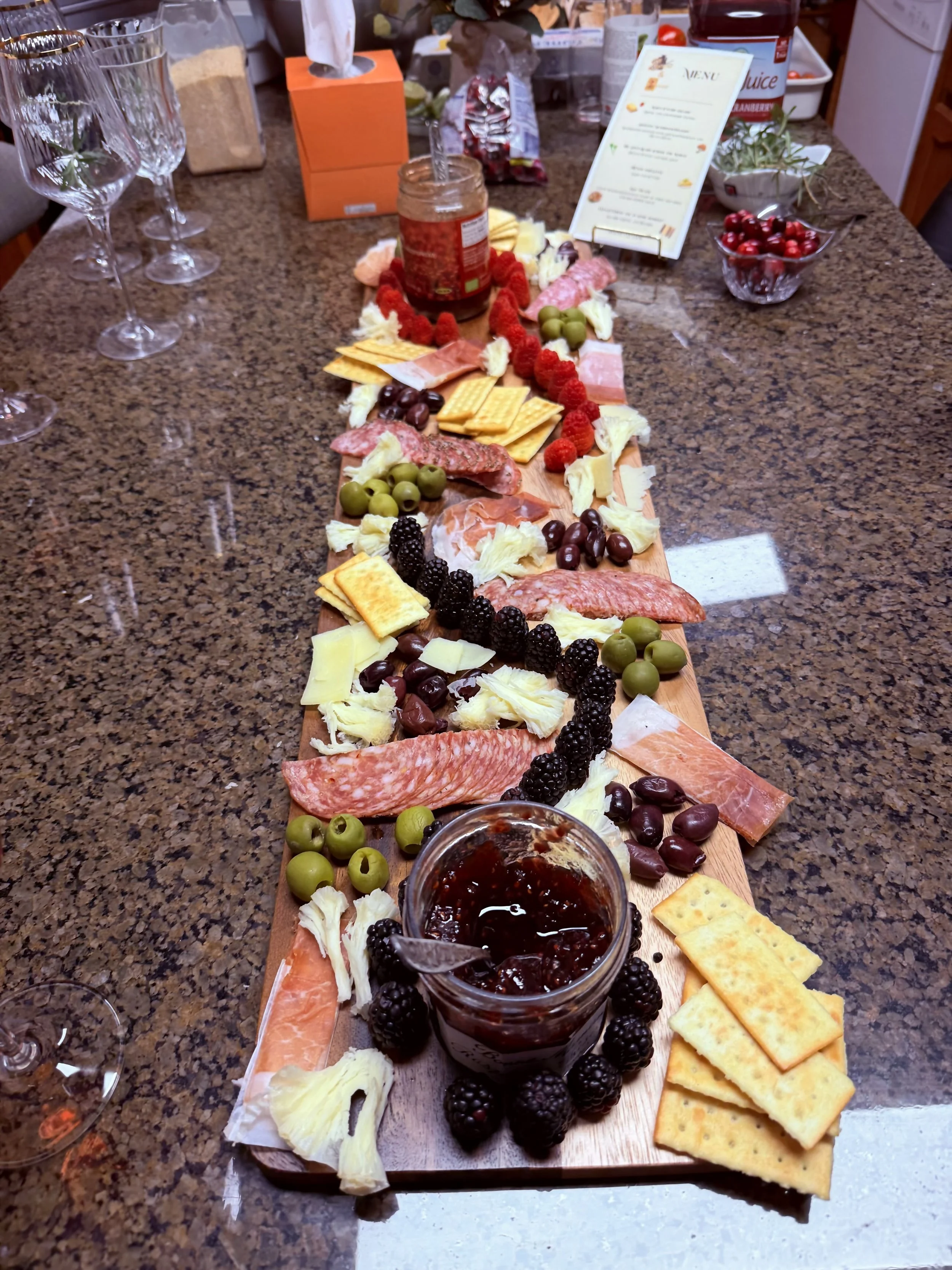 A charcuterie board with cheeses, meats, grapes, blackberries, and crackers on a wooden platter, with a jar of jam in the foreground on a granite countertop.
