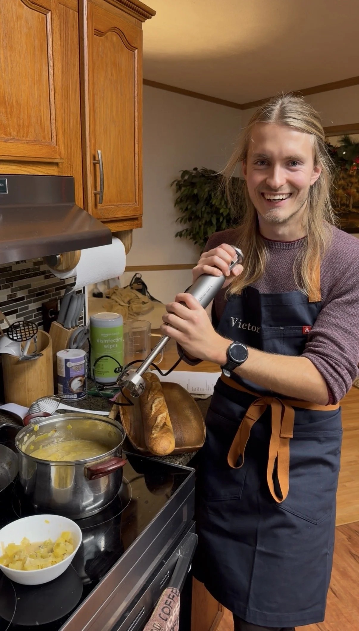 A man with long blonde hair, wearing a gray long-sleeve shirt, a black apron with the name Victor, and a wristwatch, is smiling and holding an immersion blender over a dish in a kitchen. The kitchen has wooden cabinets, a stove with a pot of yellow soup, and various kitchen items on the counter.