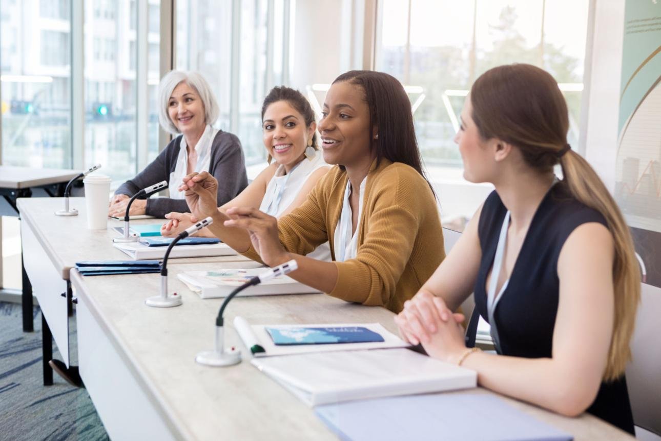 Four women sitting at a conference table with microphones, engaged in a discussion, in a bright, modern office with large windows.