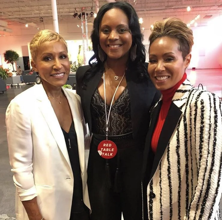 Three women standing together at an indoor event, smiling at the camera. The middle woman is wearing a black lace top, black blazer, and has a red badge that reads 'Red Table Talk'. The woman on the left is wearing a white blazer, and the woman on the right is wearing a black blazer with black and white striped pattern and a red turtleneck underneath.