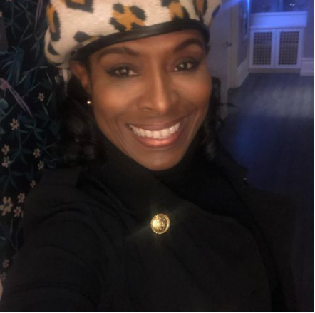 A woman smiling at the camera, wearing a leopard print hat and a black jacket with gold buttons, indoors in a well-lit room.
