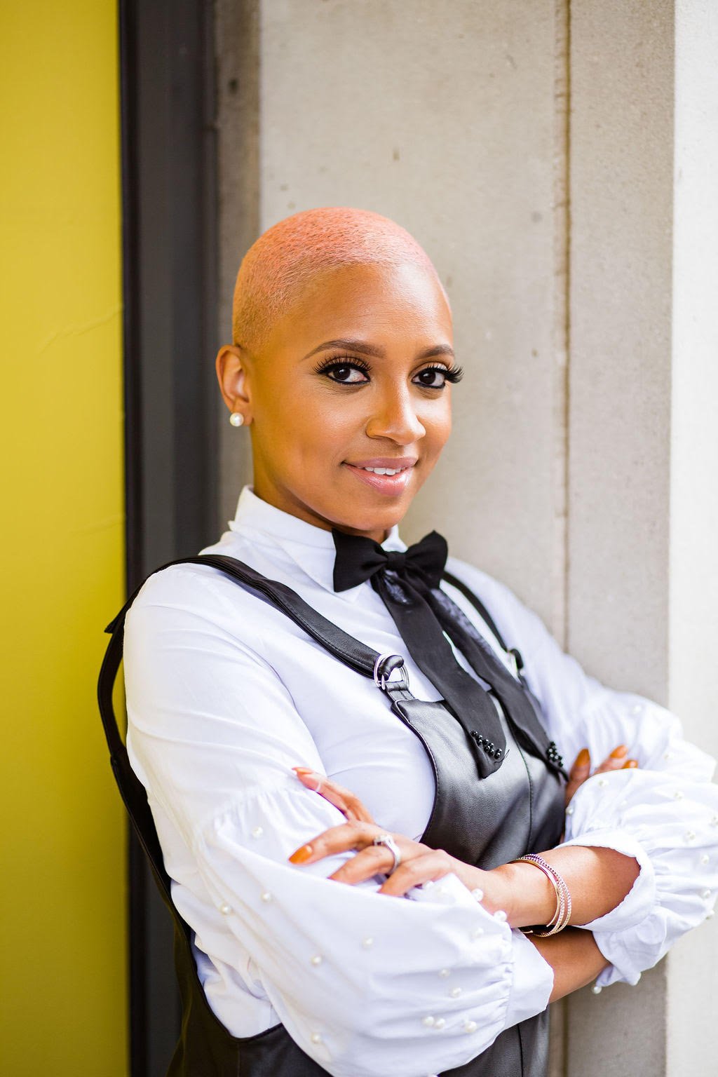 A woman with a shaved head and earrings, wearing a white top with pearl accents, a black bow tie, and a black apron, standing with arms crossed and smiling.