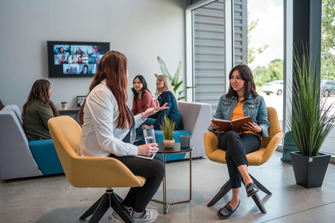Two women having a conversation in an office lounge, with several women sitting in the background, a large screen on the wall showing a video call, and large windows letting in natural light.