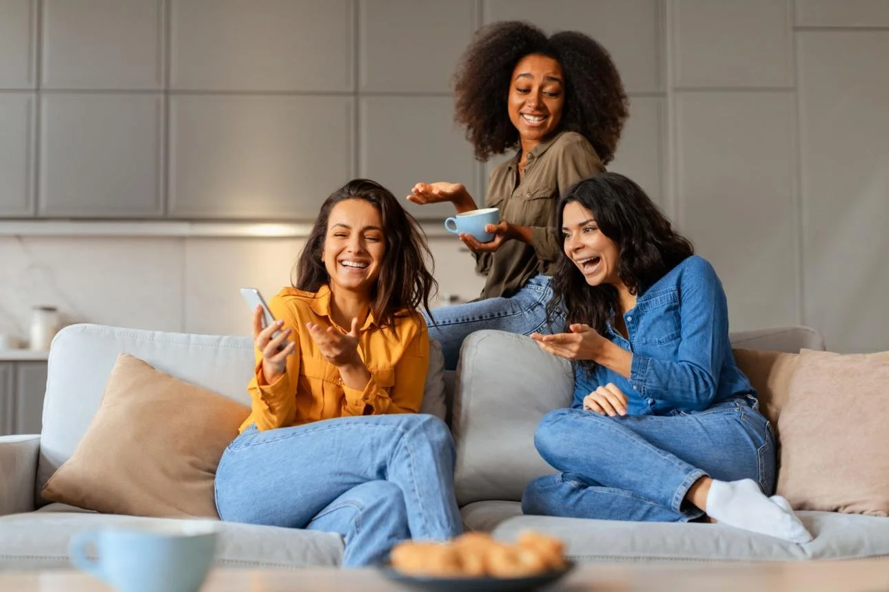 Three women sitting on a sofa, laughing and talking, with one holding a smartphone, in a cozy living room.
