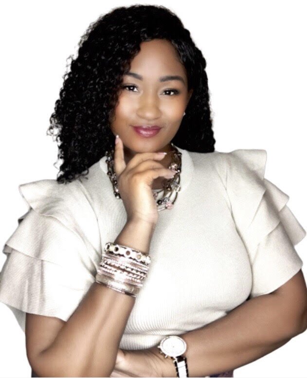 A woman with curly black hair, wearing a cream-colored top with ruffled sleeves, jewelry including bracelets, a necklace, and a watch, poses with her chin resting on her hand against a white background.