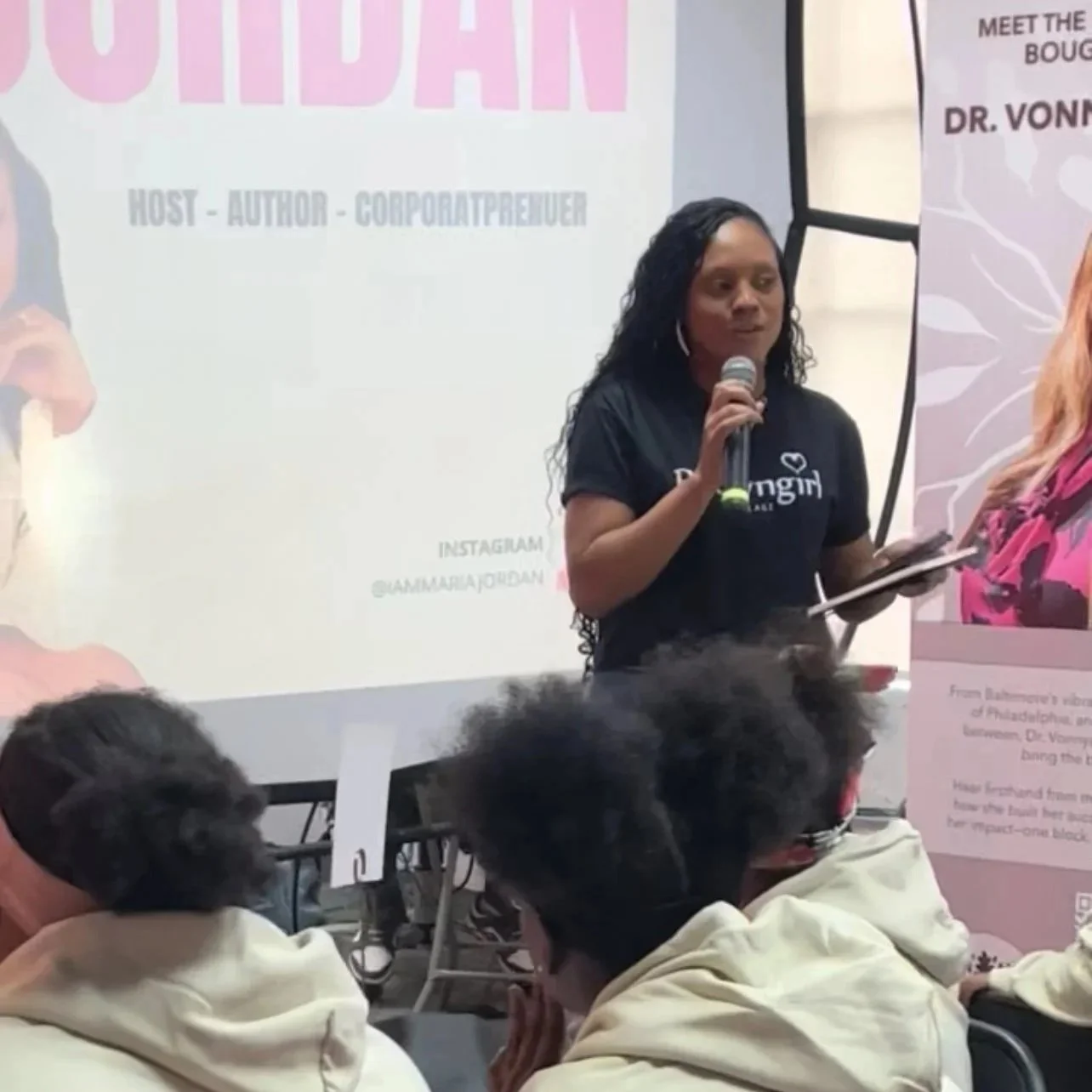 A woman speaking into a microphone at a presentation in front of an audience. There is a projection screen with text behind her, and a banner with a woman's photograph to her right.