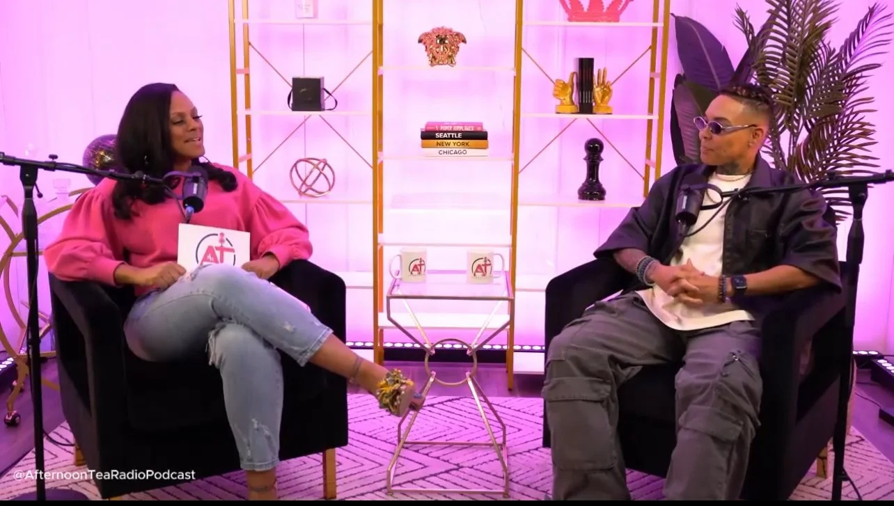 Two people seated in black armchairs on a pink-lit set, engaged in conversation with microphones, behind a white and gold shelving unit decorated with books and accessories, and a large plant on the right.