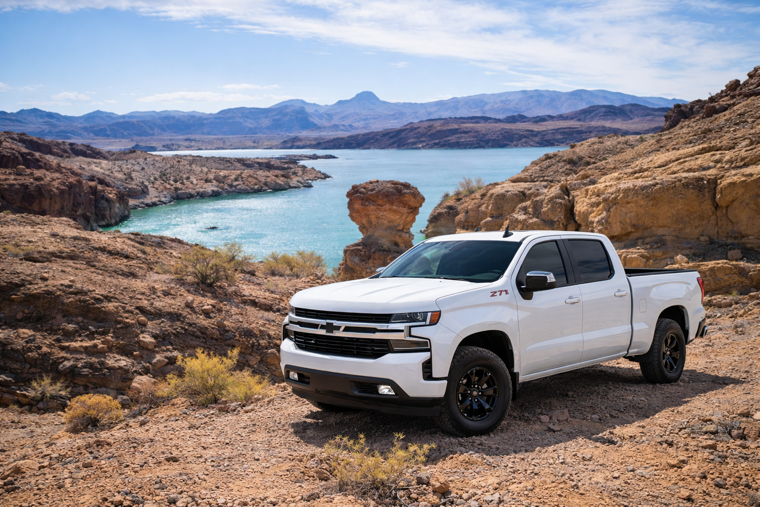 A white Chevrolet pickup truck parked on rocky terrain near a lake with mountains in the background.