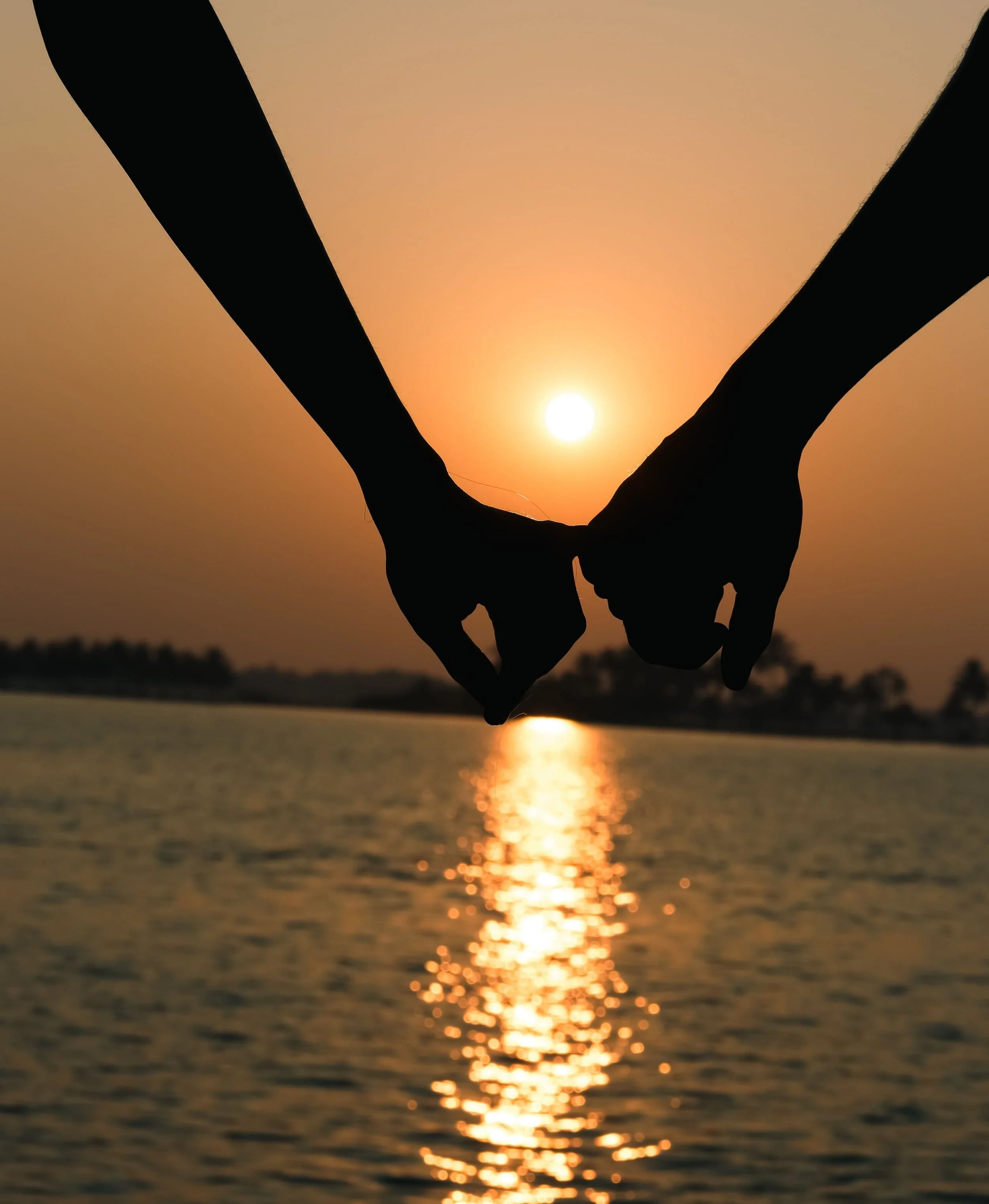 Silhouette of two hands holding each other with a sunset over a body of water in the background.