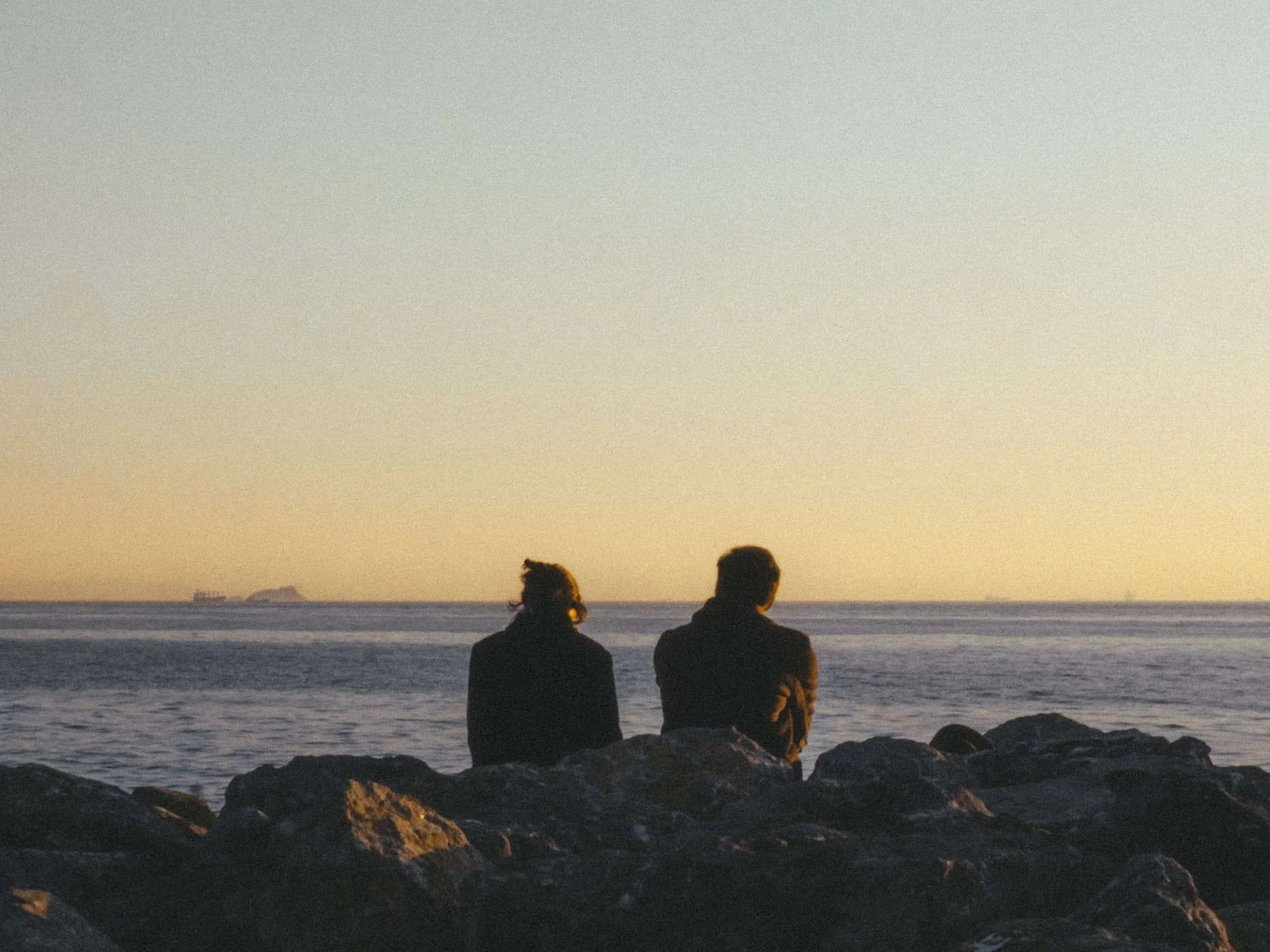 A man and woman sitting on rocks in front of the ocean at sunset.