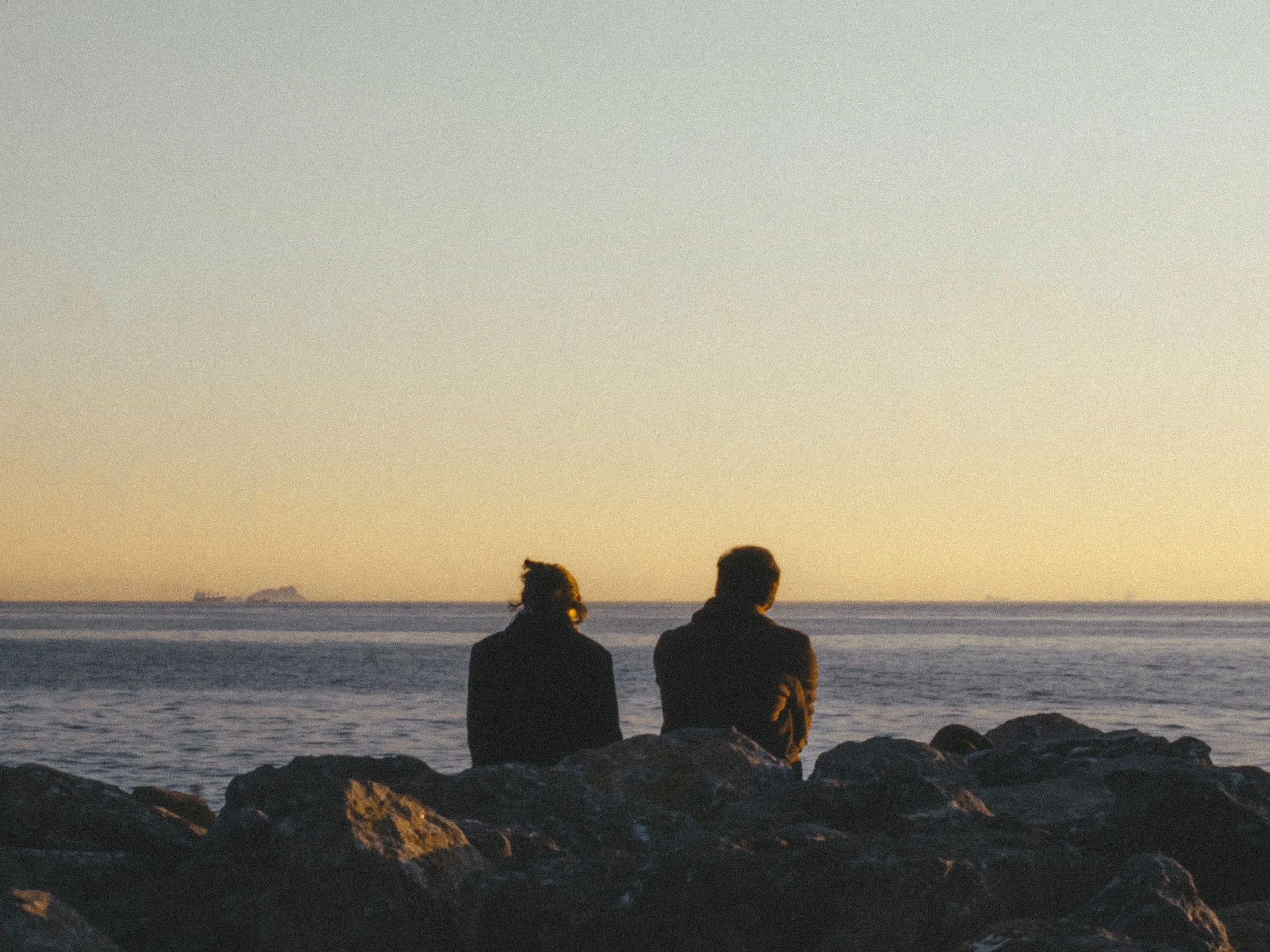 Two people sitting on rocks by the ocean at sunset, facing the water and a distant ship on the horizon.