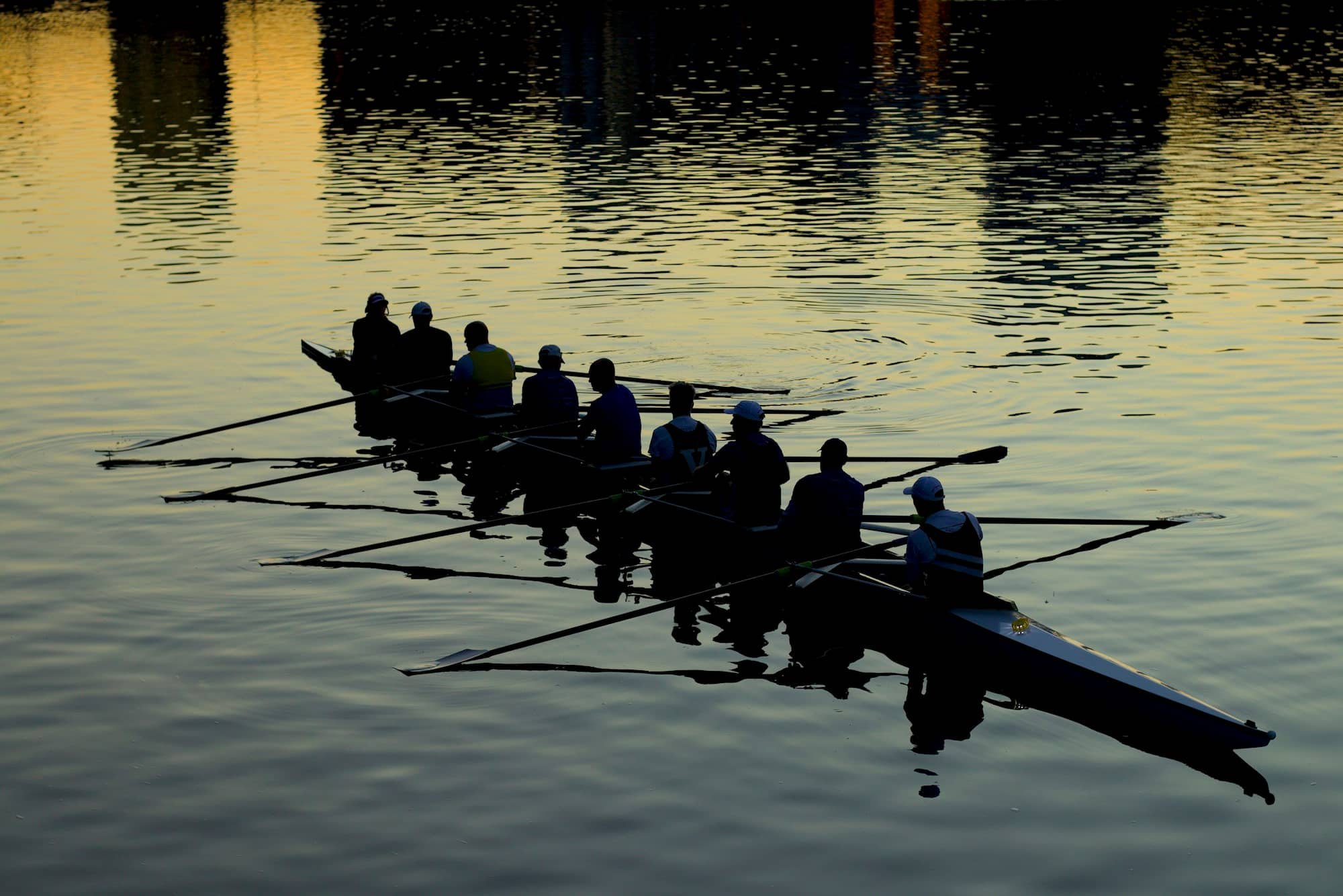 Silhouettes of people rowing a boat on calm water during sunset, with the reflections of buildings on the water.