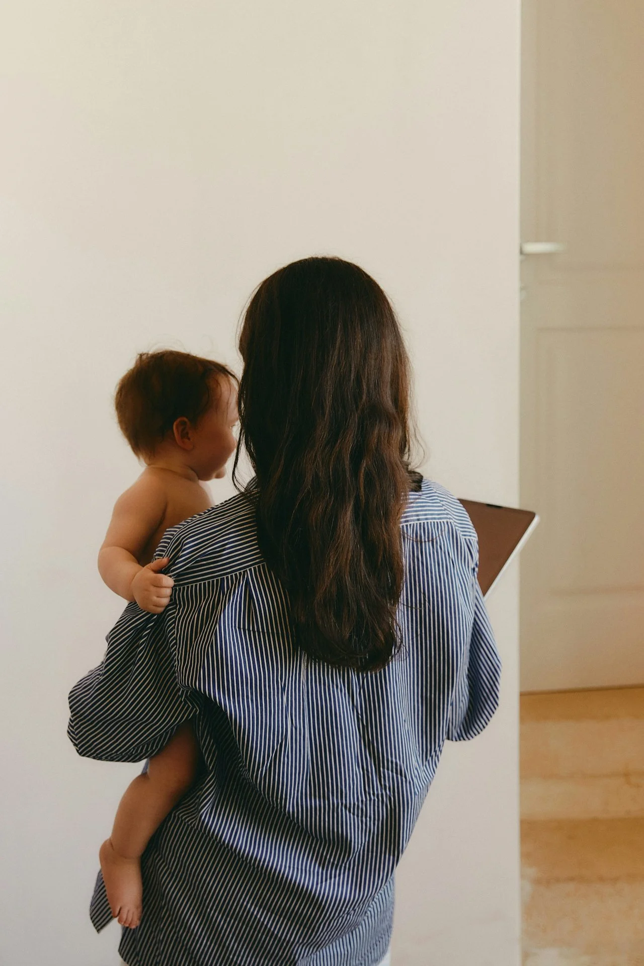 A woman with long brown hair holding a smiling toddler on her shoulder, both facing away from the camera, inside a room with white walls.