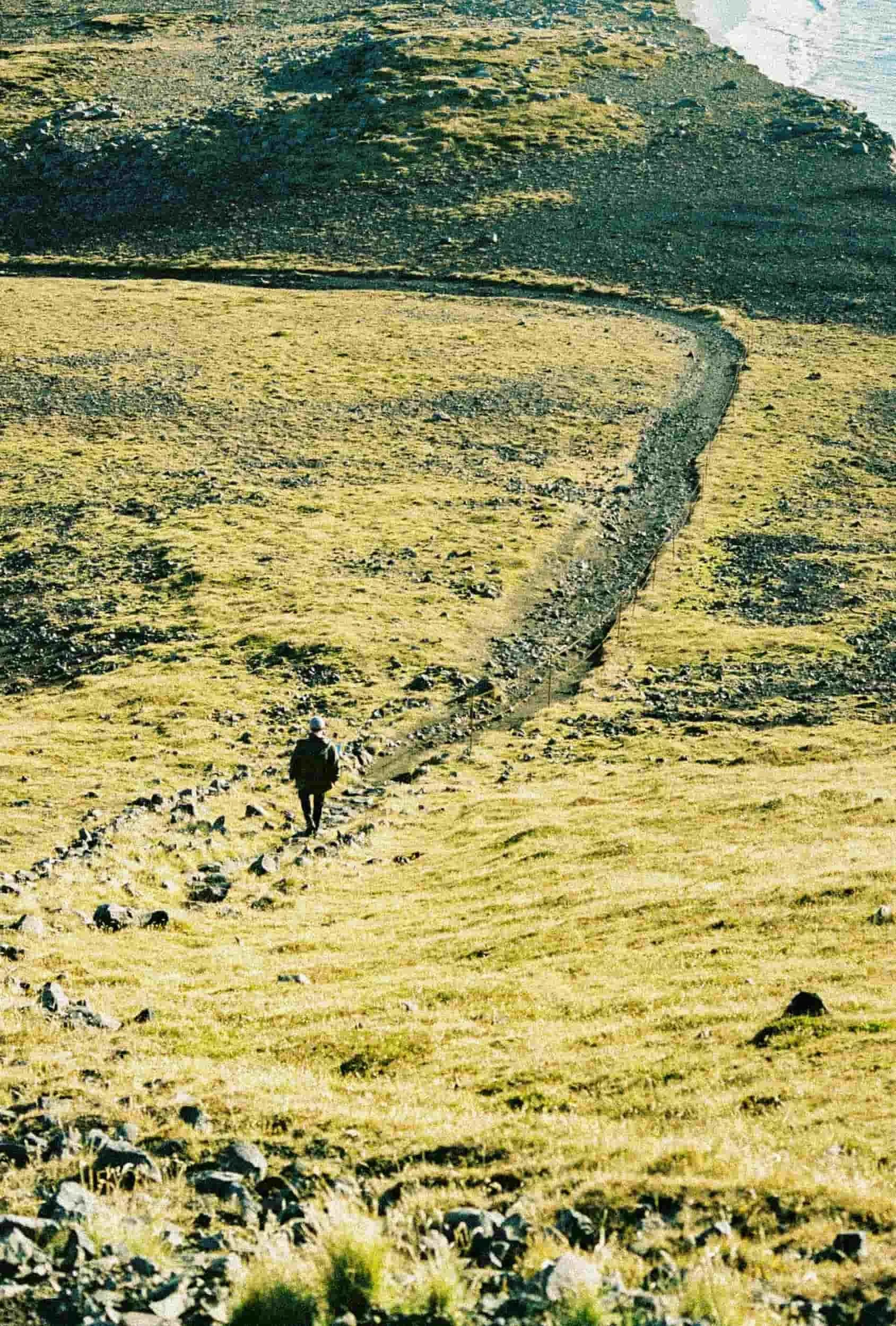 A person walking on a dirt path through a grassy, hilly landscape with large rock formations and a cliff in the background.