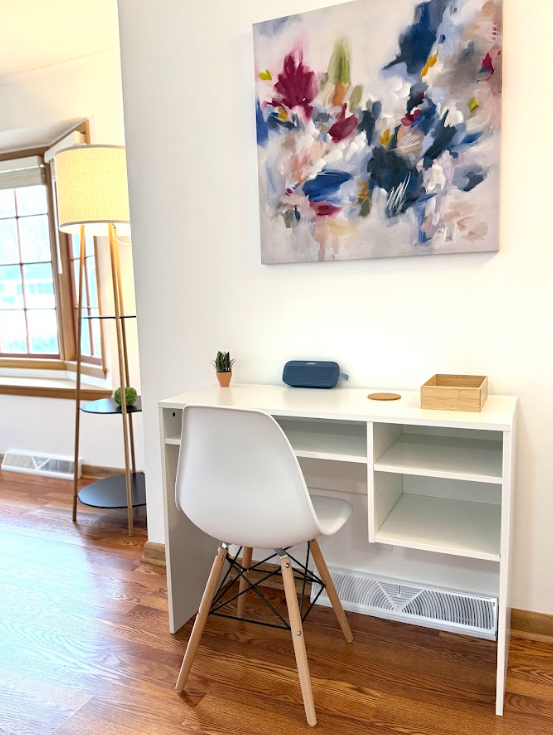 White desk with an empty shelf on the right and a small potted cactus on top, a white modern chair, a colorful abstract painting hanging on the wall, and a tall floor lamp beside a window in a bright room.