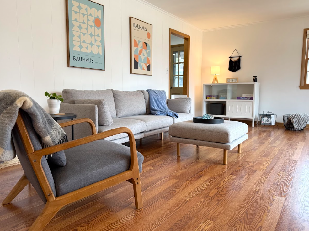 Living room with a gray sofa, a wooden armchair with gray cushions, a round ottoman, wall art, a white cabinet, a table lamp, and decorative items on the furniture and floor, all on wooden floors.