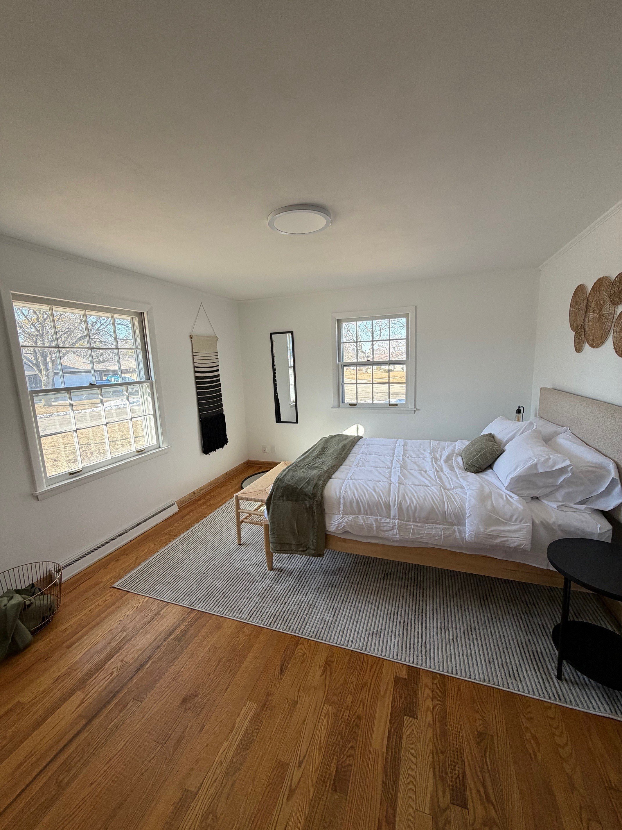 A cozy bedroom with hardwood floors, a large window, white walls, and a bed with white bedding and green pillows. There is a small round black side table, a striped rug, and wall decor including three woven circular art pieces and two narrow mirrors. Sunlight is streaming through the windows, illuminating the room.