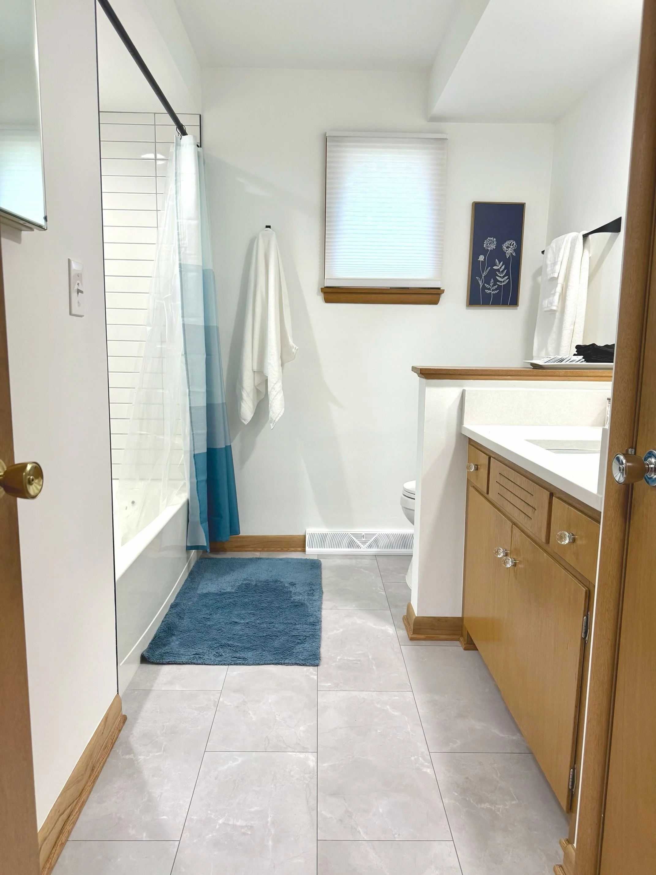 A bathroom with beige tile floor, white walls, a window with closed blinds, a blue rug, a white and blue shower curtain, a white towel hanging on a black towel bar, a framed botanical art print, a wooden vanity with a white countertop, and a vent on the floor.