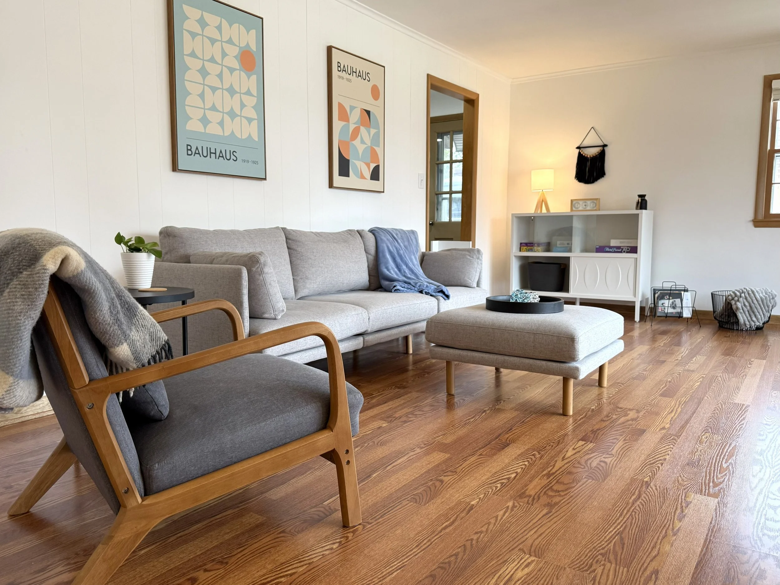 A cozy living room with a light gray sofa adorned with a blue blanket, a round black coffee table, a white bookshelf with various items, a table lamp, wall art featuring geometric designs labeled 'BAUHAUS 1919-1925', a stool, an armchair with a throw blanket, and a black wall hanging. The room has natural wood floors and trim, with windows providing natural light.