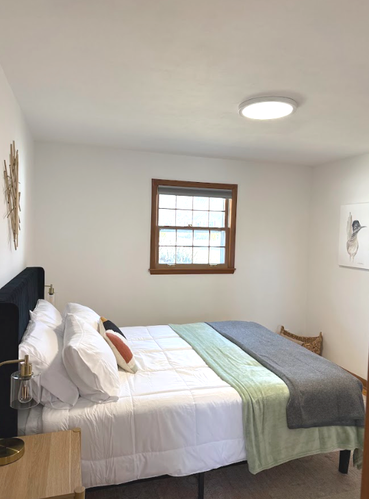 A neatly made bed with white bedding and pillows, a small nightstand, and a window with a wooden frame in a minimalist bedroom with white walls and a ceiling light.