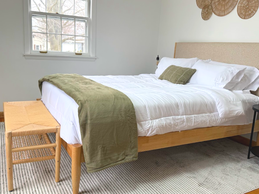 A neatly made bed with white bedding, a green blanket, and an olive green pillow in a bright bedroom. There is a wooden stool at the foot of the bed, a small black side table, a window with white trim, and decorative wall art above the bed.