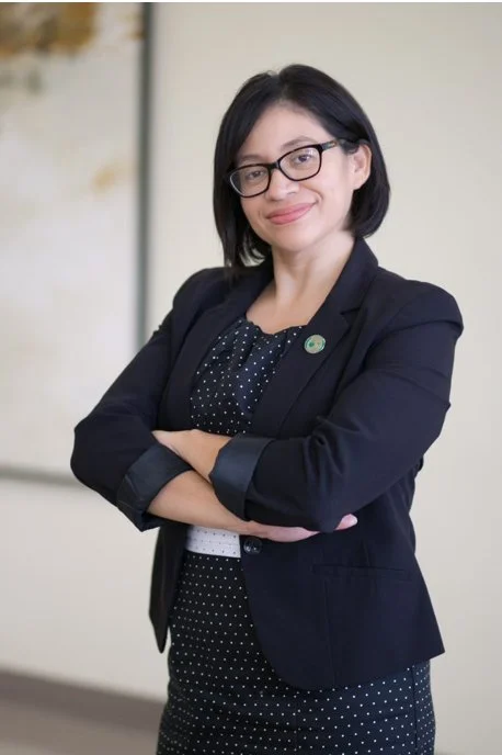 A woman with shoulder-length black hair, glasses, and a confident smile, dressed in a dark blazer and polka dot dress, standing with arms crossed in an office or professional setting.