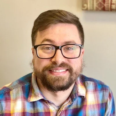 Close-up of a man with glasses and a beard smiling, wearing a plaid shirt, in an indoor setting.
