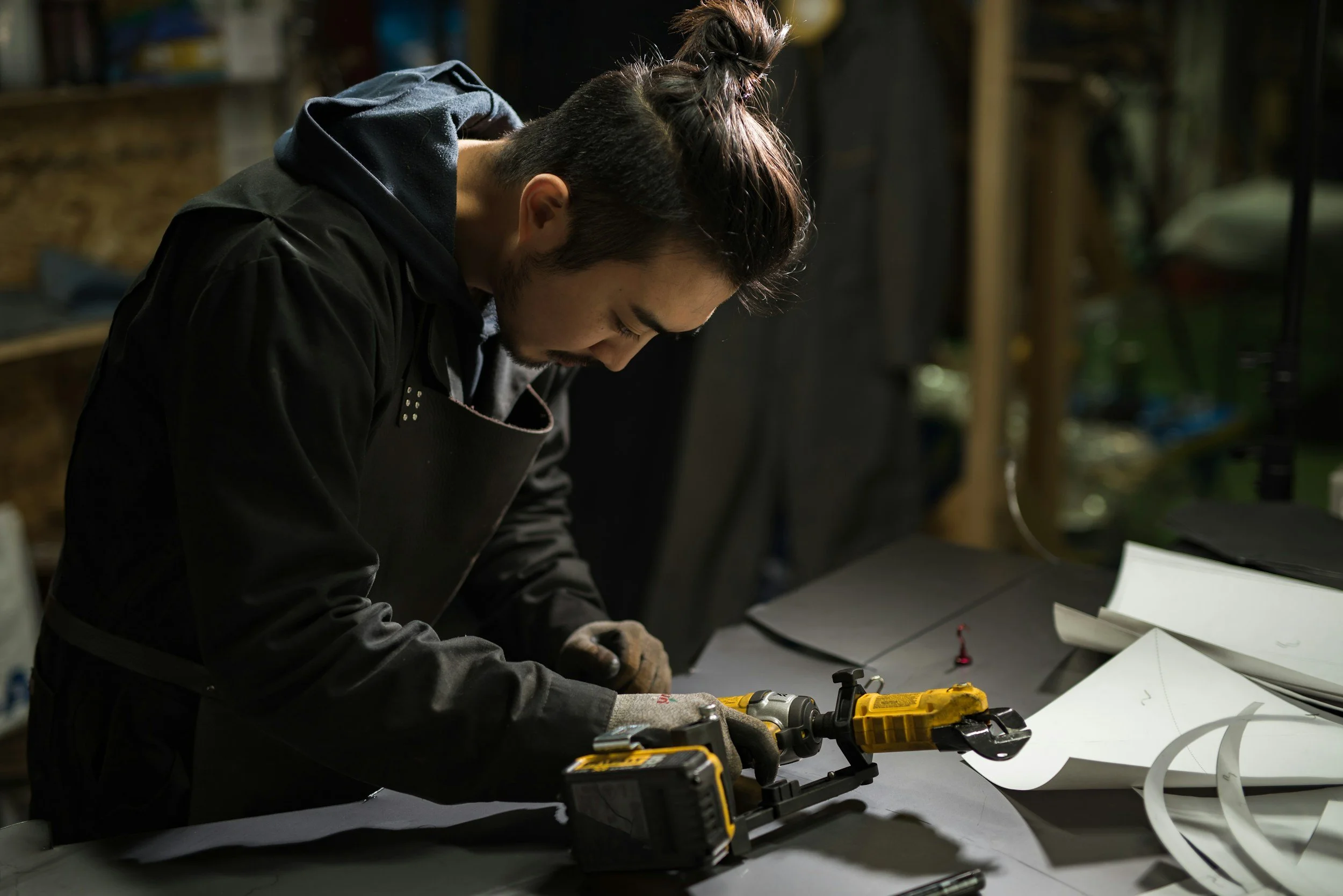 Man working with construction tools in a workshop, wearing gloves and concentrating on a project, surrounded by papers and equipment.