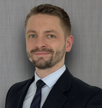 A man with light skin, short brown hair, and a beard, dressed in a dark suit, white shirt, and striped tie, standing against a gray background.