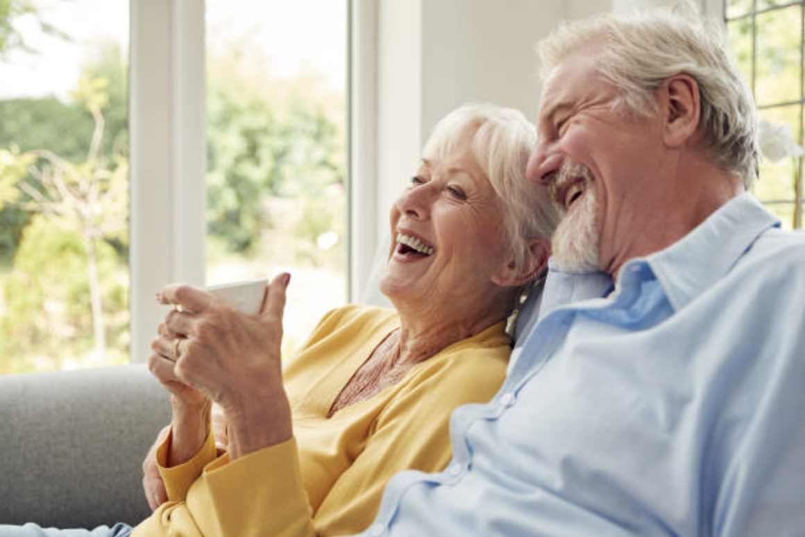 An elderly woman and man sitting on a sofa indoors, laughing and sharing a joyful moment while looking at a cup.