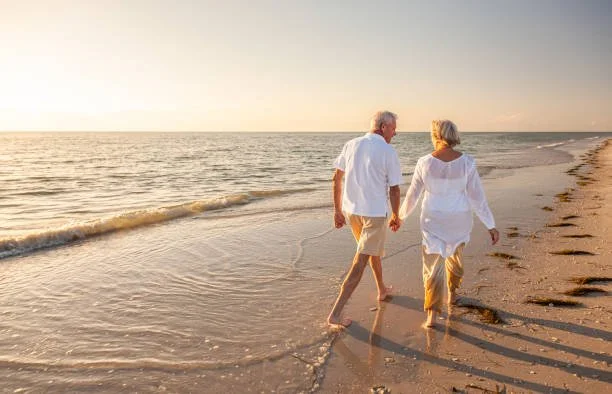 Older couple walking hand in hand on a sandy beach at sunset.