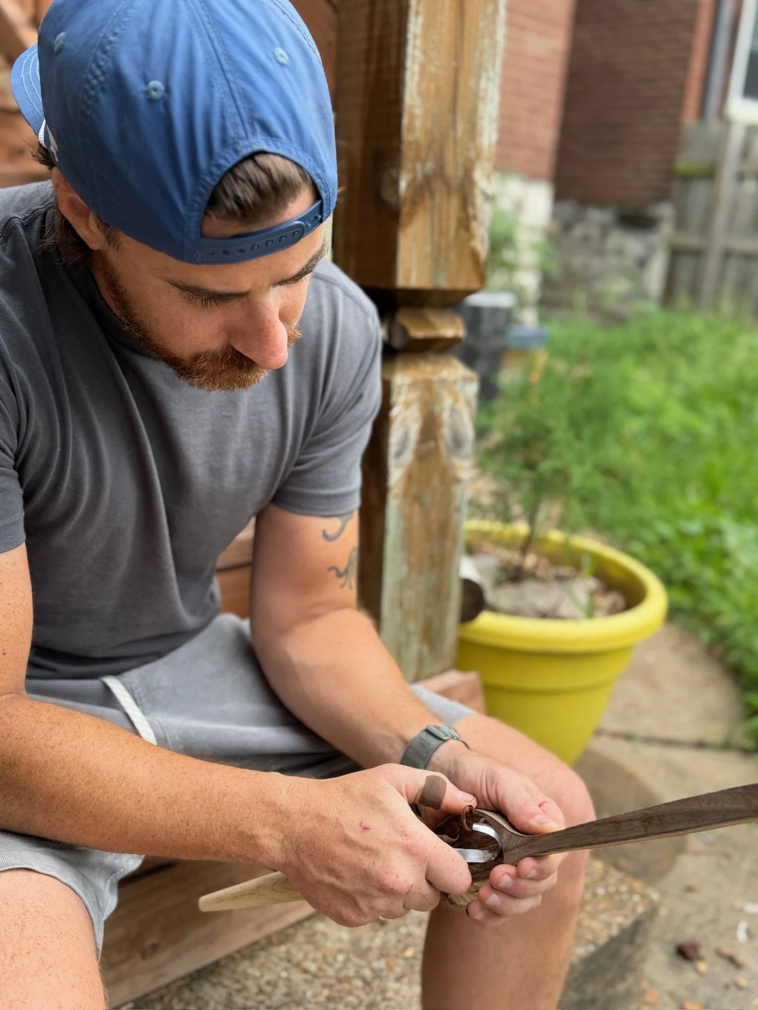 A man wearing a blue baseball cap, gray t-shirt, and gray shorts is using a hand saw to cut a piece of wood outdoors, with a wooden structure and a yellow plant pot in the background.