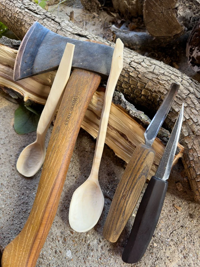 A collection of woodworking tools including an axe, a chisel, a carving knife, and two wooden spoons on a dirt surface, with chopped logs in the background.