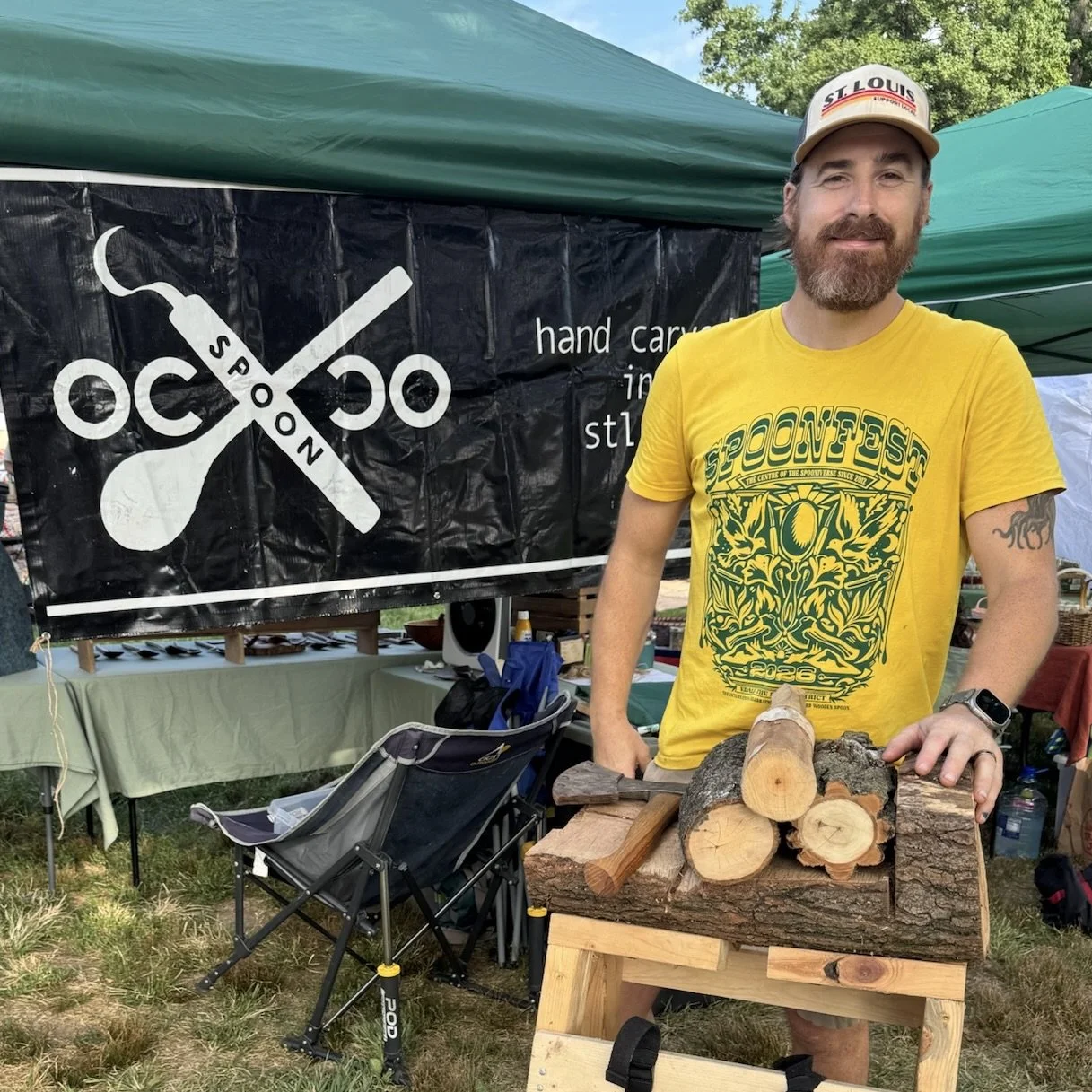 A man with a beard and a yellow T-shirt standing outdoors at a booth, with logs on a wooden stand in front of him and a black banner behind with scissors and the words "OCO SPOON" on it.