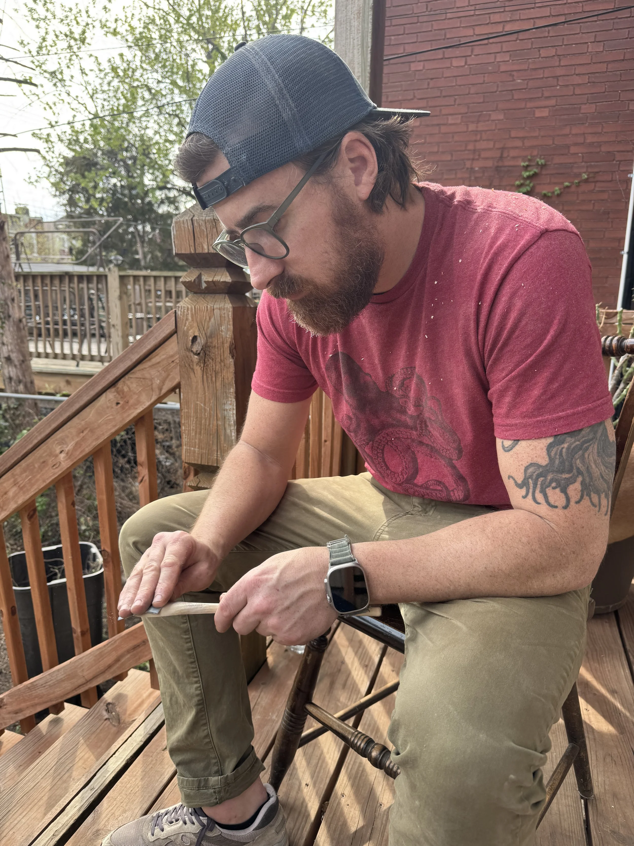 A man with a beard, glasses, and a backwards baseball cap sitting on a wooden deck. He is wearing a red T-shirt with a design, beige pants, sneakers, and an Apple Watch, looking down and holding an object in his hands. There are trees, a brick wall, and some outdoor furniture in the background.