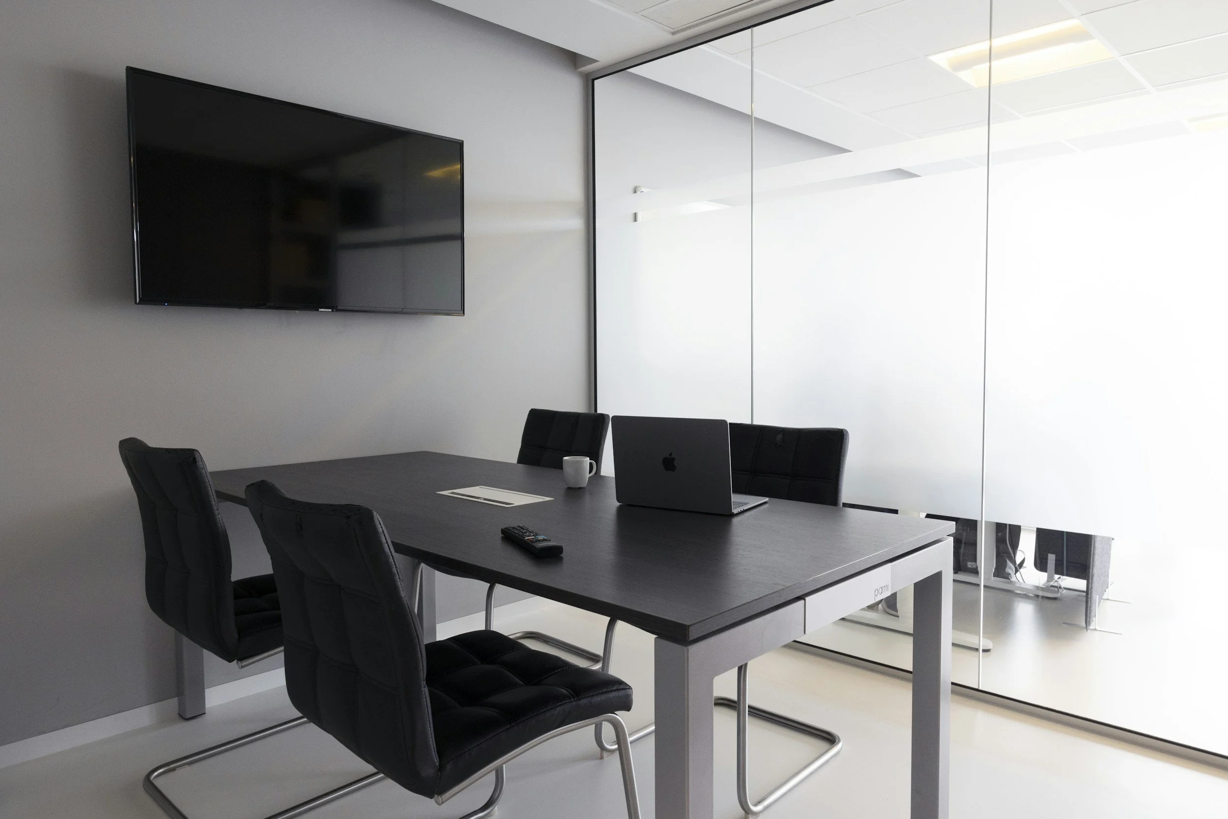 Modern conference room with a black table, four black chairs, a wall-mounted television, a laptop, a coffee mug, a remote, and a glass wall with frosted sections.