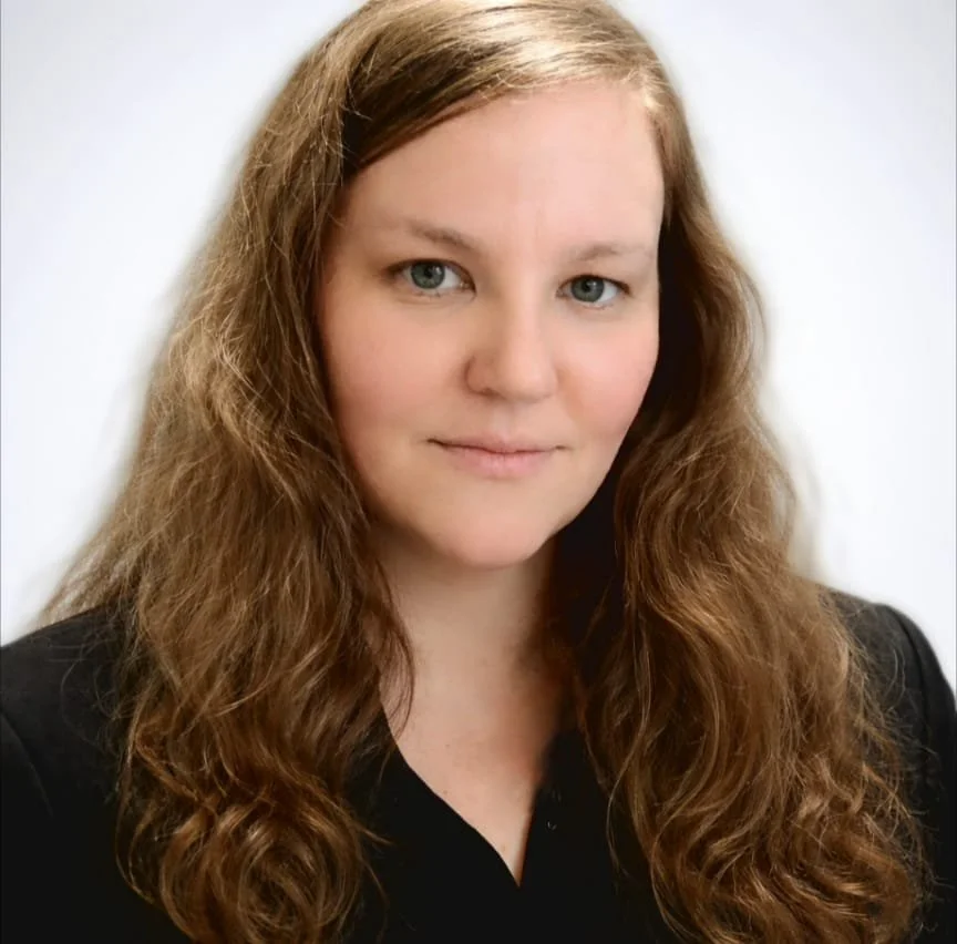 A woman with long, wavy red hair and blue eyes looking at the camera with a slight smile, wearing a black top against a white background.