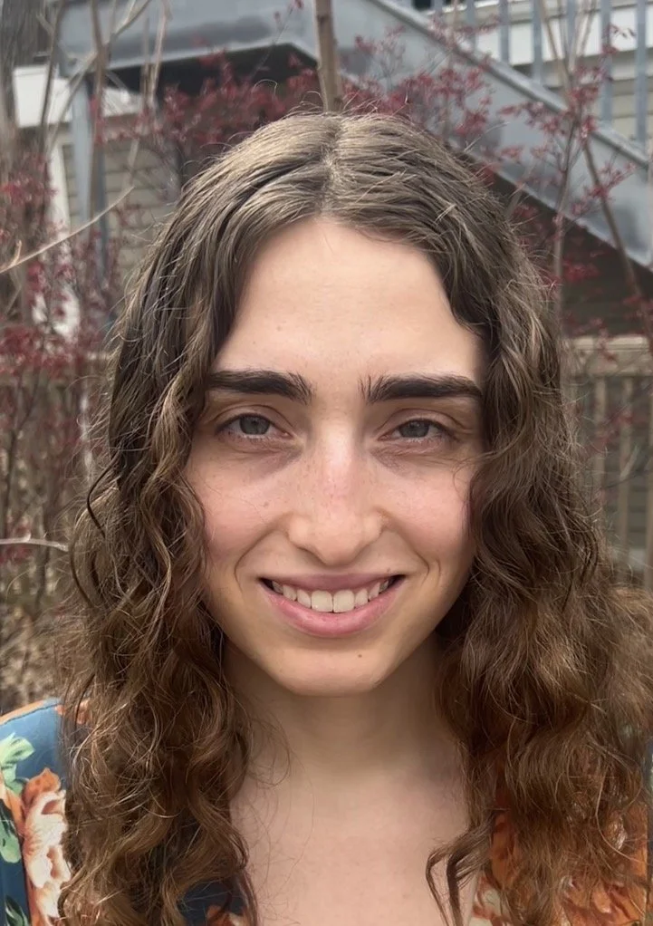 A young woman with long, curly brown hair smiling outdoors with trees and a building in the background.