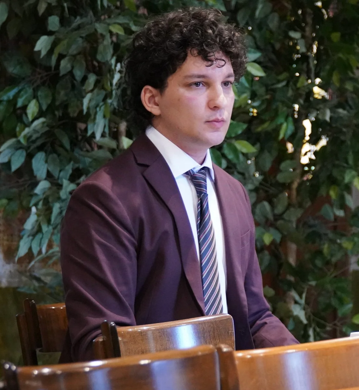 A young man with curly dark hair wearing a burgundy suit jacket, white shirt, and striped tie, sitting indoors with green leafy plants in the background.