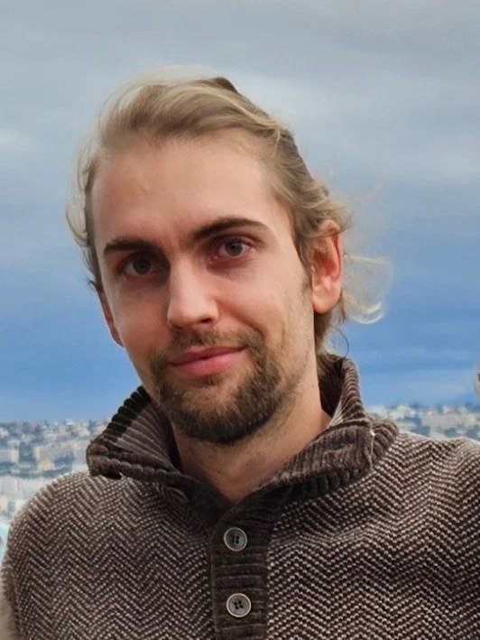 Young man with blond hair and a beard, wearing a brown sweater, standing outdoors with cityscape and cloudy sky in the background.