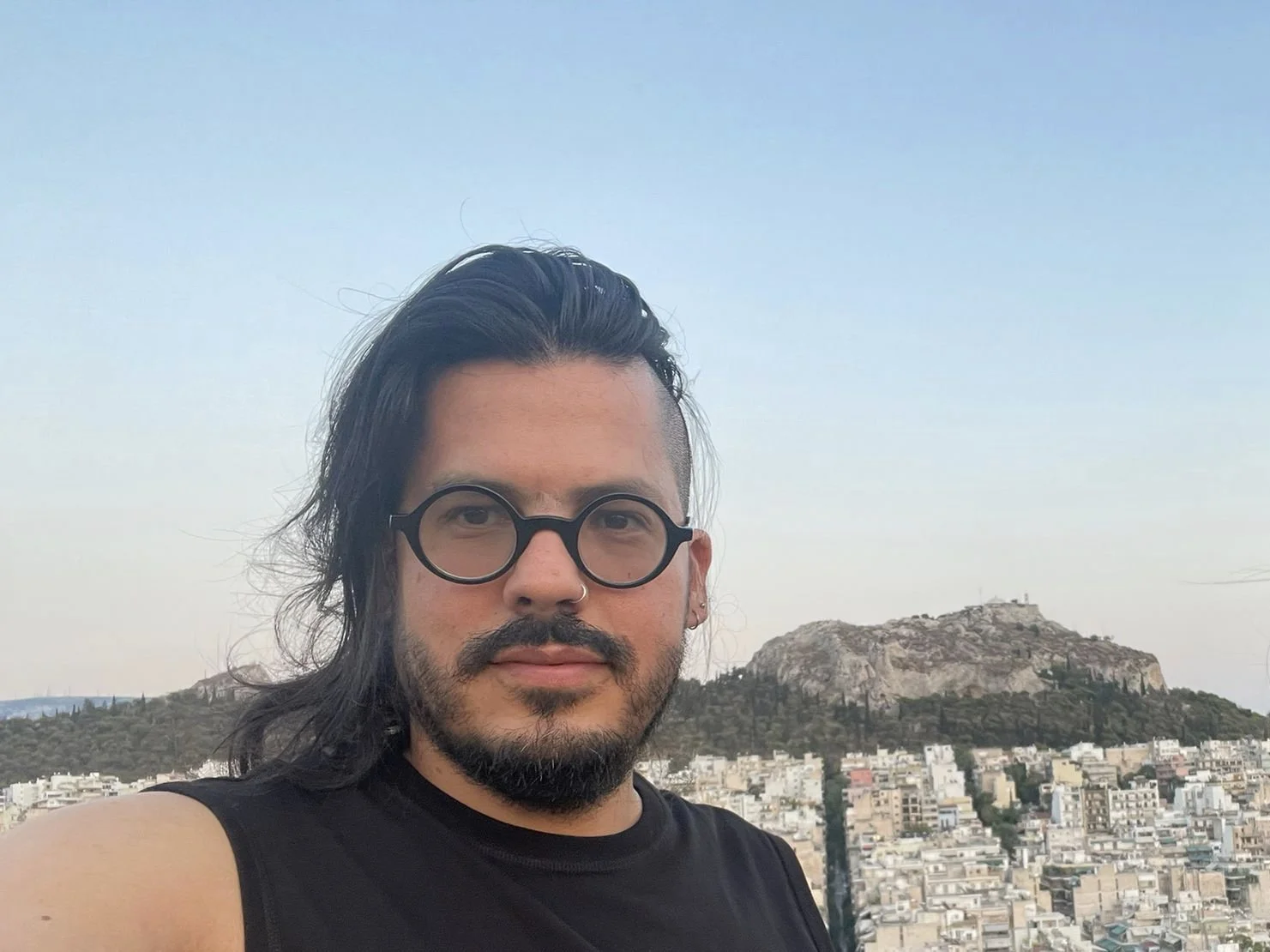 A man with black hair, a beard, round glasses, and a nose ring taking a selfie with a cityscape and a hilltop in the background.