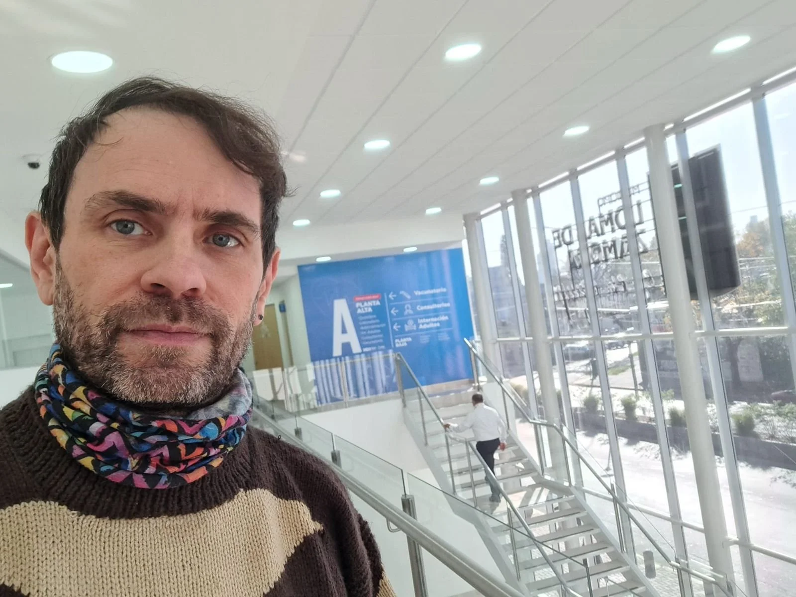 A man with a beard and light-colored eyes taking a selfie inside an airport. The airport has large glass windows, and a staircase with a person ascending. There are signs and placement of advertisement boards in the background.