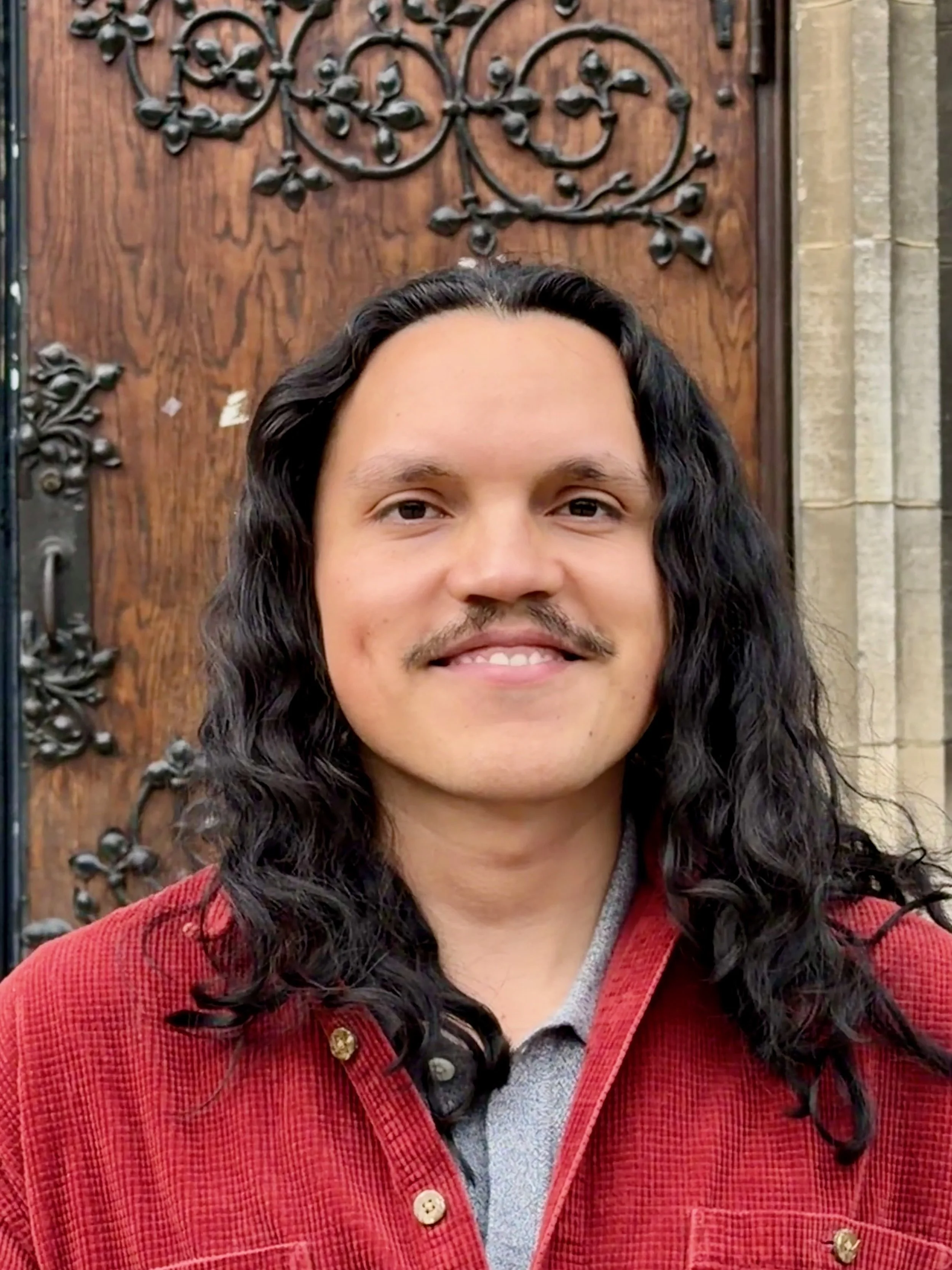 Close-up of a smiling man with long curly dark hair standing in front of a decorated wooden door.