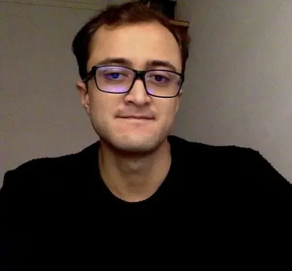 A young man with light skin, short brown hair, wearing glasses with a dark frame, and a black shirt. He is indoors in front of a plain, grayish wall.