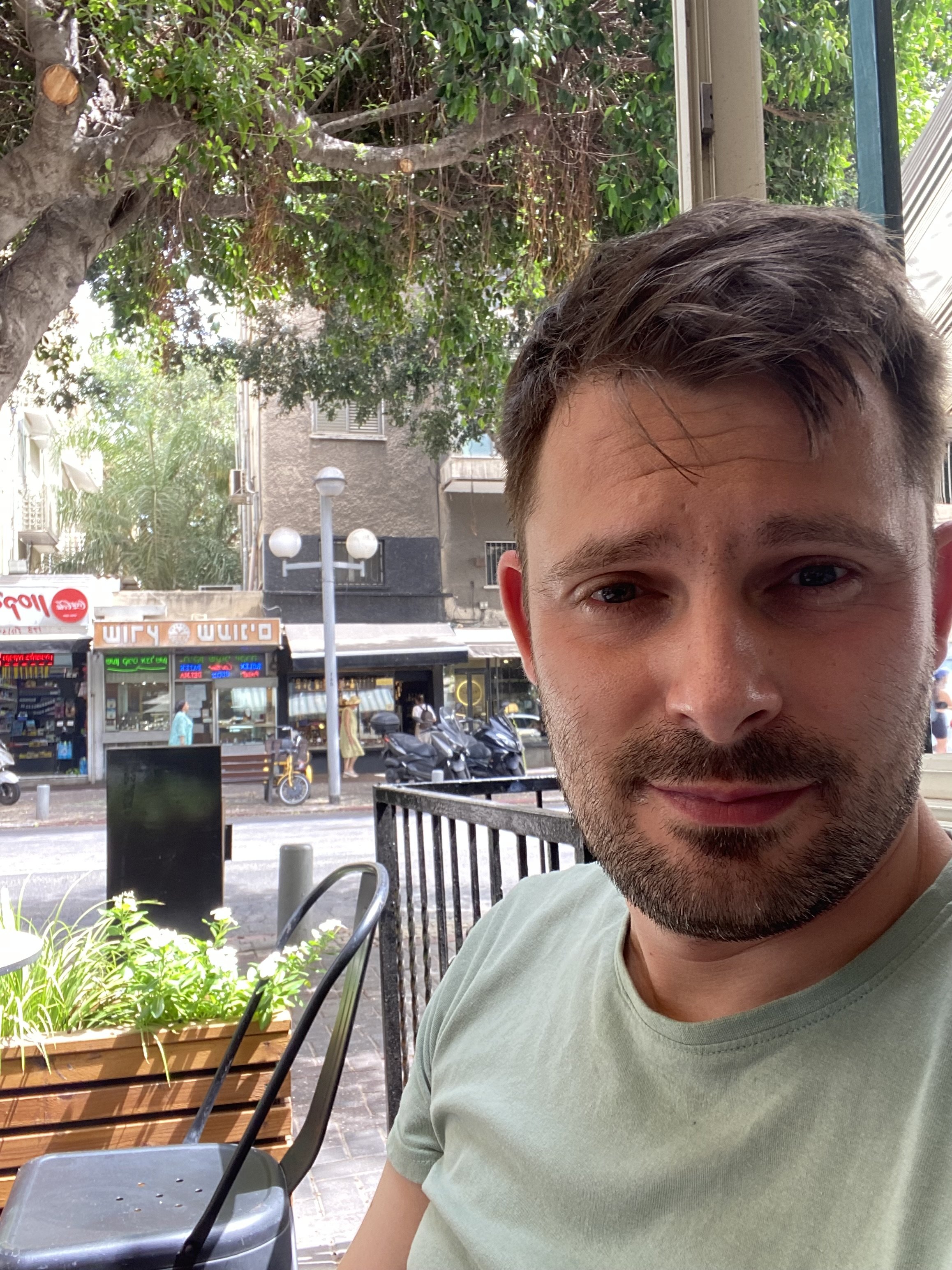 Close-up of a man with brown hair, stubble, and a light green t-shirt sitting outdoors at a café with trees, parked scooters, and a storefront in the background.