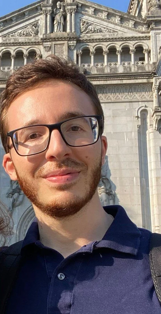 A young man with glasses and a beard taking a selfie outdoors in front of a historic building with ornate stone carvings and statues.