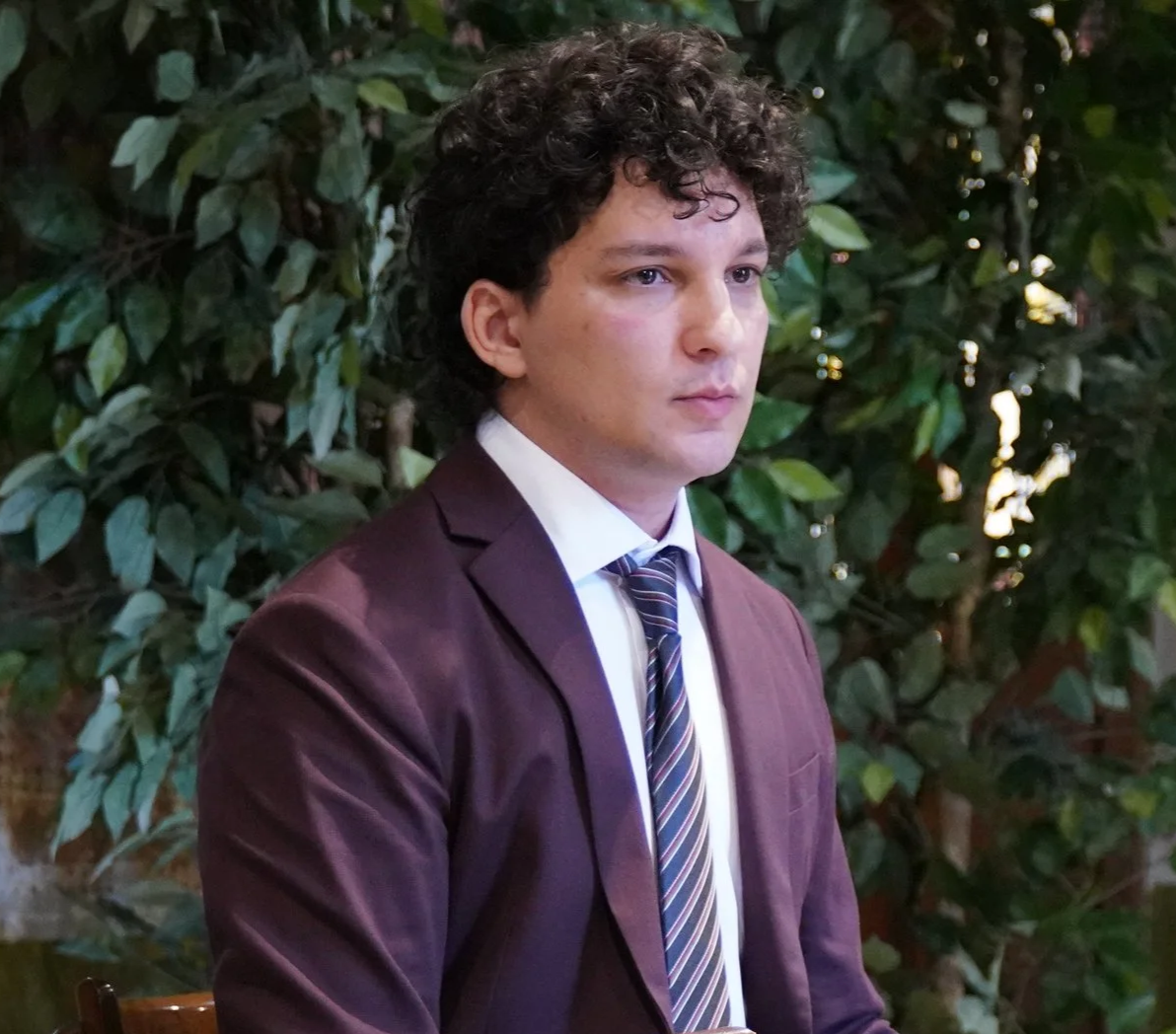 A young man with curly dark hair and light skin, wearing a burgundy suit, white dress shirt, and striped tie, sitting outdoors in front of green leafy bushes.