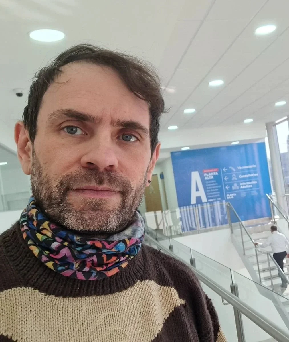 A man with brown hair, blue eyes, and a beard taking a selfie at an airport. He is wearing a patterned neck gaiter and a brown and cream sweater. Behind him, there is a large blue sign with information about entrances and services, and airport staff are visible.